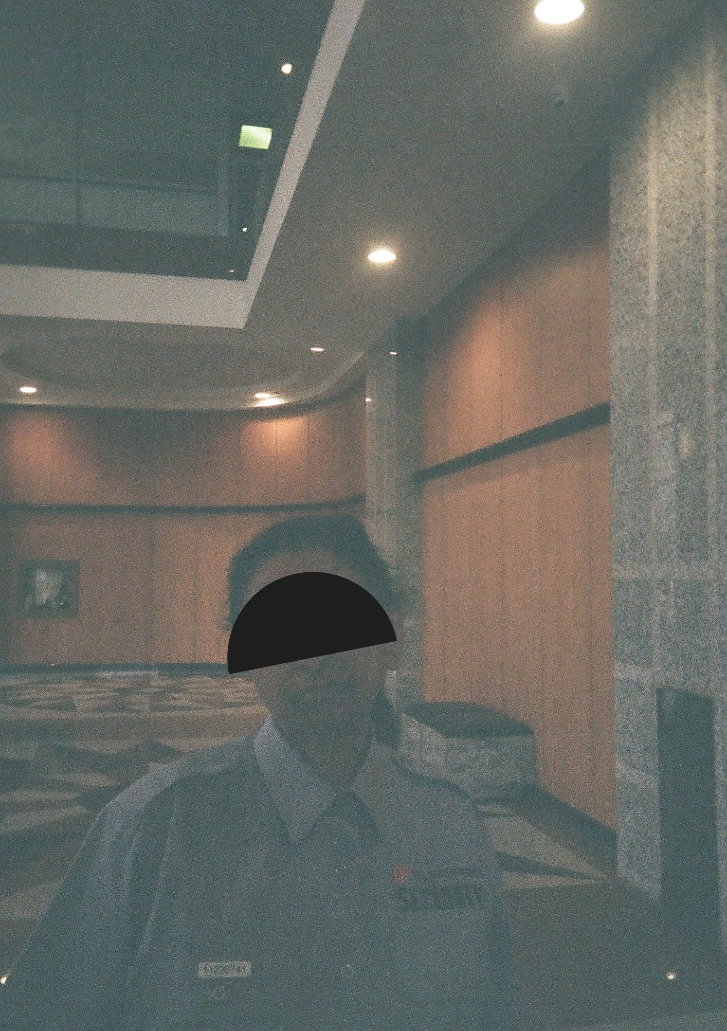 Grainy photo of a grimacing security guard. The guard's face is obcured by a black semicircle, tilted on an angle.