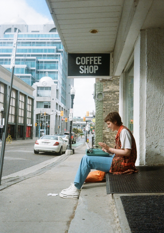 Sadie sitting on a sidewalk-facing stoop, typewriter in lap, with the corner clock face of 55 King St W visible in the backgorund.